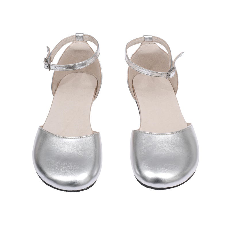 Puede incluir: Un par de bailarinas plateadas de punta cerrada con correas en el tobillo. Los zapatos tienen un brillo met&aacute;lico y un peque&ntilde;o cierre de hebilla. Las suelas son negras y el interior es de color beige claro. Los zapatos est&aacute;n dise&ntilde;ados para una ocasi&oacute;n formal o elegante.