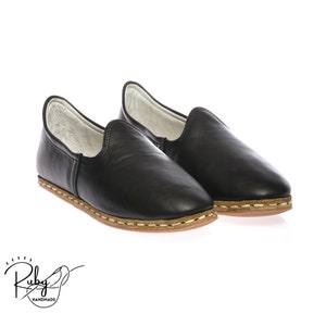 Op de afbeelding: Een paar zwarte leren slippers met een bruine gestikte rand en een witte fleece voering.