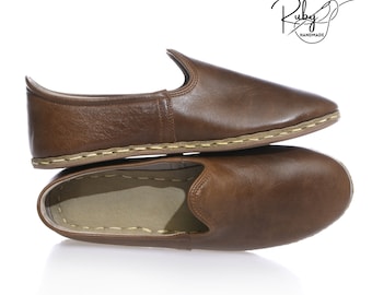 Op de afbeelding: Een paar bruine leren slippers met een gestikte naad rond de rand. De slippers liggen op elkaar gestapeld. De merknaam "Ruby Handmade" is zichtbaar in de rechterbovenhoek van de afbeelding.