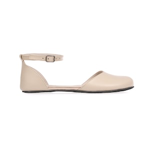 May include: A beige flat sandal with an ankle strap and buckle closure. The shoe has a rounded toe and a low, black sole. The sandal is made of a smooth material and is set against a white background.