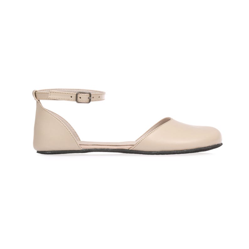 May include: A beige flat sandal with an ankle strap and buckle closure. The shoe has a rounded toe and a low, black sole. The sandal is made of a smooth material and is set against a white background.