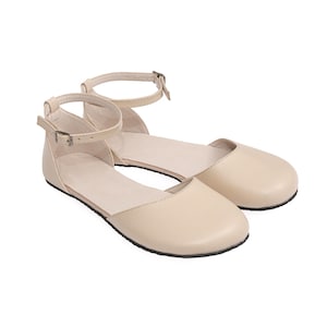 May include: A pair of beige leather flats with ankle straps. The shoes have a rounded toe design and a small buckle closure on the ankle strap. The soles are black.
