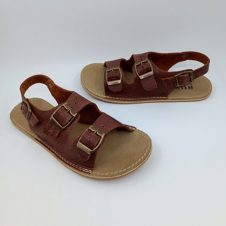 May include: A pair of brown leather sandals with two adjustable buckles on each strap. The sandals have a back strap and a beige sole.