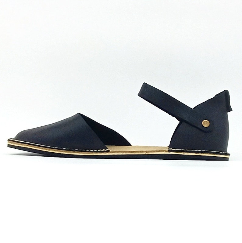 Puede incluir: Sandalias de cuero negro con una correa de tobillo y un detalle de bot&oacute;n dorado. Las sandalias tienen una suela plana y una punta puntiaguda.