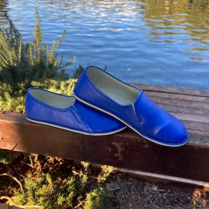 Op de afbeelding: Een paar blauwe leren instapschoenen met witte stiksels. De schoenen liggen op een houten oppervlak.