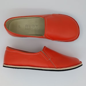 May include: A pair of bright orange leather slip-on shoes with a white sole and the text "RUBY BAREFOOT" on the inside of one shoe.