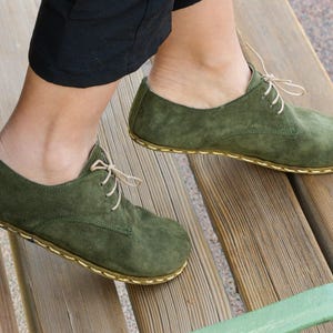 Op de afbeelding: Olijfgroene suède schoenen met beige veters en stiksels. De schoenen hebben een klassieke derby-stijl met een ronde neus. De afbeelding toont de schoenen op een houten bank, wat wijst op een buitenomgeving. De schoenen lijken ongedragen.