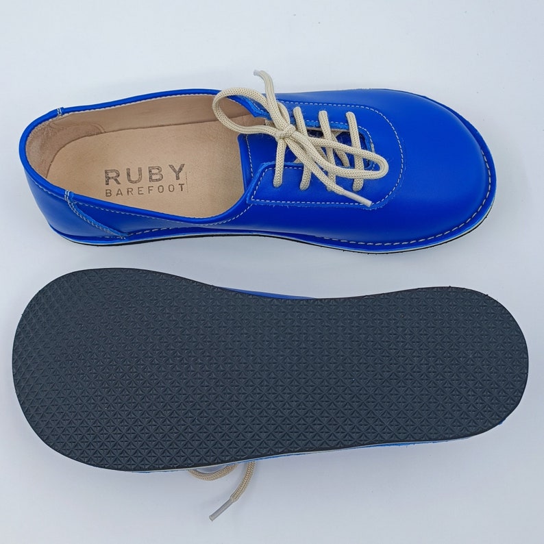 May include: Blue leather lace-up shoes with a white sole and a black rubber outsole. The shoes have a label that reads "RUBY BAREFOOT".