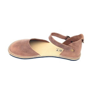 May include: Brown leather sandals with an ankle strap and buckle closure. The sandals have a simple design and are made of high-quality leather. The brand name "BY EFOOT" is printed on the side of the sandal.