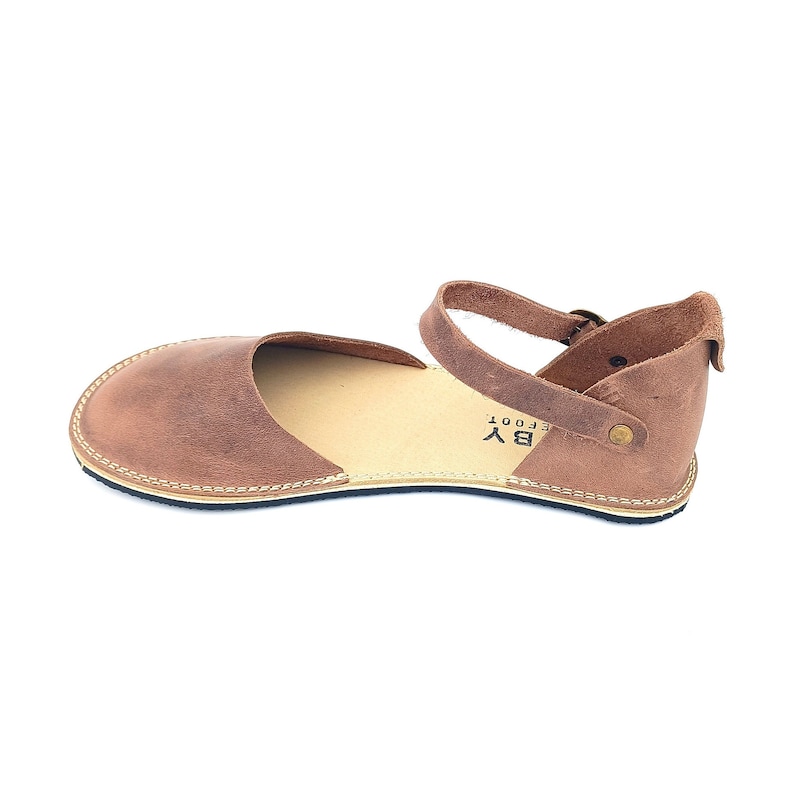 May include: Brown leather sandals with an ankle strap and buckle closure. The sandals have a simple design and are made of high-quality leather. The brand name "BY EFOOT" is printed on the side of the sandal.