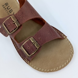 May include: Brown leather sandals with two adjustable buckles. The sandals have a stitched edge and a brown leather sole.