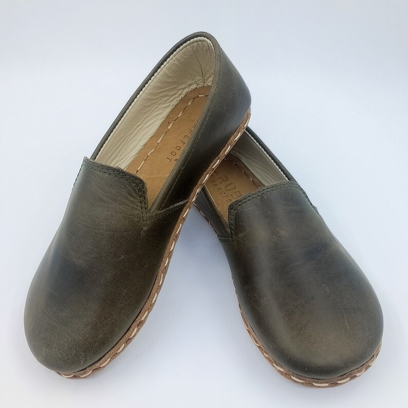 K&ouml;nnte beinhalten: Ein Paar dunkelgr&uuml;ne Slipper aus Leder mit wei&szlig;en N&auml;hten an der Sohle. Die Schuhe haben einen leicht abgenutzten Look und sind aus weichem, geschmeidigem Leder gefertigt.