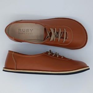 May include: A pair of brown leather lace-up shoes with a white sole. The shoes have the text "RUBY BAREFOOT" on the inside of the shoe.