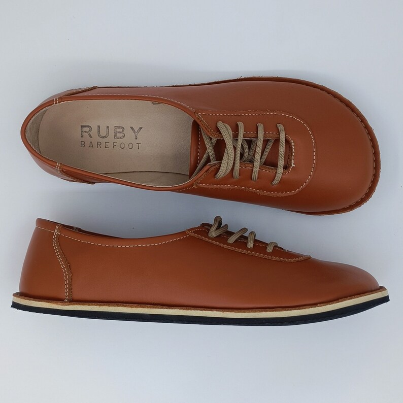 May include: A pair of brown leather lace-up shoes with a white sole. The shoes have the text "RUBY BAREFOOT" on the inside of the shoe.