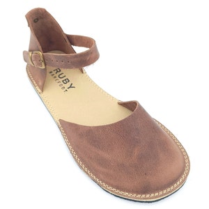 May include: Brown leather ankle strap sandals with a buckle closure. The sandals have a rounded toe and a flat sole. The brand name "RUBY BAREFOOT" is printed on the insole.