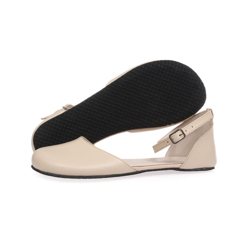 May include: Cream-colored flats with an ankle strap and buckle closure. The shoes have a rounded toe and a textured black sole. The design features a cutout on the side of the shoe.
