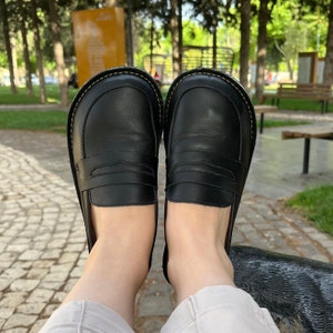 May include: A pair of black leather loafers with a simple design. The loafers have a rounded toe and a low heel. The shoes are worn by a person sitting on a bench.