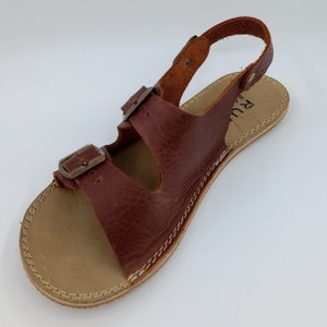 May include: Brown leather sandals with two buckles and an ankle strap. The sandals have a tan leather sole with stitching around the edge.