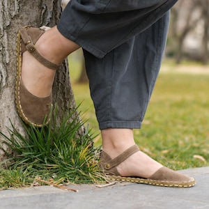 Pode incluir: Sandálias de camurça castanhas com tira e fecho de fivela. As sandálias têm bico arredondado e são costuradas com linha contrastante. As solas são rasas e as sandálias são usadas ao ar livre.