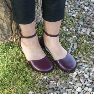 Op de afbeelding: Bordeauxkleurige ballerina's met enkelbandjes. De schoenen hebben een ronde neus en een lage hak. De afbeelding toont de schoenen op een grasachtige ondergrond met kleine stenen en een boomstam op de achtergrond. De schoenen worden gedragen met een zwarte broek.