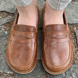 Può includere: Un paio di mocassini in pelle marrone con un classico design penny loafer. Le scarpe hanno una punta arrotondata e una suola cucita. La pelle ha un aspetto strutturato. Le scarpe sono indossate su una superficie di pietra.