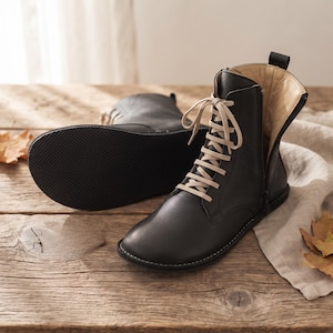 Puede incluir: Un par de botines de cuero negro con cordones beige, cierre de cremallera lateral y suela de goma negra texturizada. El forro interior es de color beige claro y tiene una pequeña lengüeta negra en la parte superior del talón.
