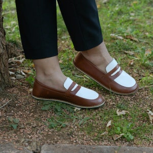 May include: Brown leather loafers with white leather accents. The loafers have a slip-on design and a rounded toe.