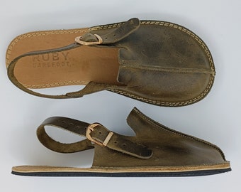 Puede incluir: Un par de sandalias de cuero verde oliva con cierre de hebilla y plantilla de cuero marrón. Las sandalias tienen un logotipo "RUBY BAREFOOT" en la plantilla.