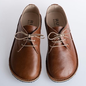 Puede incluir: Un par de zapatos marrones de cuero con cordones, suela blanca y banda de rodadura texturizada. Los zapatos tienen un diseño sencillo y están hechos de cuero de alta calidad.