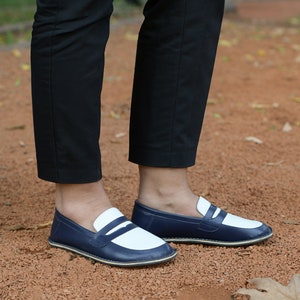 May include: A pair of blue and white leather loafers with a white stripe across the toe. The loafers are worn by a person standing on a red gravel surface.
