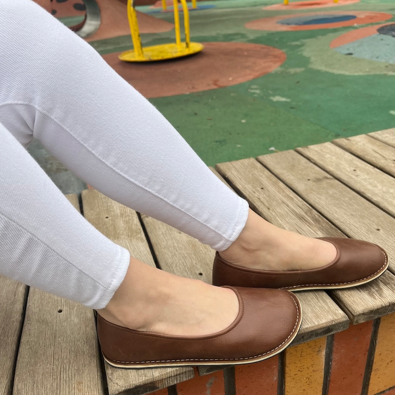 Peut inclure: Ballerines en cuir marron avec un bout arrondi et des coutures blanches. Les ballerines sont port&eacute;es avec un pantalon blanc. Les chaussures sont sur une surface en bois.