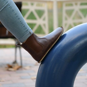 Puede incluir: Una bota de tobillo de cuero marr&oacute;n con un panel el&aacute;stico negro y una suela blanca. La bota la lleva puesta una persona y est&aacute; apoyada sobre una superficie azul.