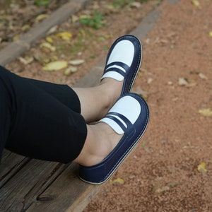 May include: A pair of blue and white loafers with a white leather upper and a blue leather trim. The loafers are worn by a person sitting on a wooden bench.