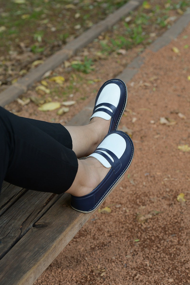 May include: A pair of blue and white loafers with a white leather upper and a blue leather trim. The loafers are worn by a person sitting on a wooden bench.