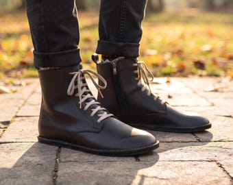 May include: A pair of black leather ankle boots featuring a smooth finish, rounded toes, and light beige laces. The boots have a side zipper closure and a flat, dark sole, worn with dark gray cuffed denim jeans in an outdoor setting.
