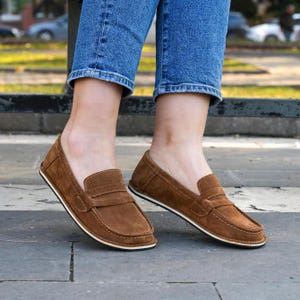 Op de afbeelding: Een paar bruine suède loafers met een witte zool. De loafers hebben een eenvoudig ontwerp met een enkele band over de bovenkant.