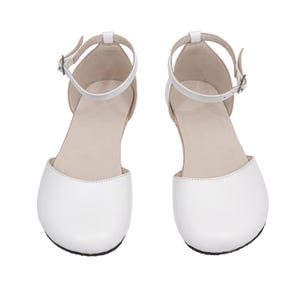 Puede incluir: Un par de sandalias blancas de punta cerrada, con correas en el tobillo y peque&ntilde;as hebillas. Los zapatos tienen una punta redondeada y una suela plana. Las sandalias est&aacute;n hechas de un material liso, probablemente similar al cuero, sobre un fondo blanco.