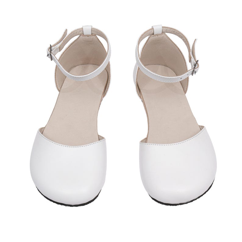 Puede incluir: Un par de sandalias blancas de punta cerrada, con correas en el tobillo y peque&ntilde;as hebillas. Los zapatos tienen una punta redondeada y una suela plana. Las sandalias est&aacute;n hechas de un material liso, probablemente similar al cuero, sobre un fondo blanco.