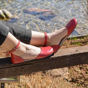 Op de afbeelding: Rode leren ballerina's met enkelbandjes. De schoenen hebben een ronde neus en een lage hak. De bandjes zijn vastgezet met kleine gespen. De schoenen worden gedragen op een houten bankje bij een water.