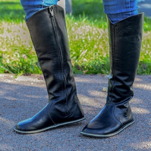May include: A pair of black knee-high boots with a side zipper. The boots are made of a smooth, shiny material and have a flat sole with a light-colored trim. The boots are worn with blue jeans.