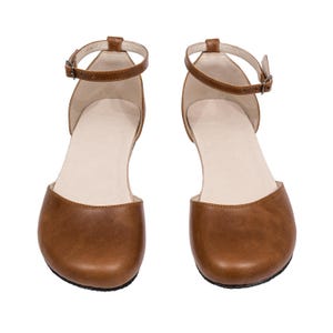 Puede incluir: Un par de sandalias de cuero marr&oacute;n con correas en el tobillo y peque&ntilde;as hebillas. Las sandalias tienen un dise&ntilde;o de punta redondeada y una plantilla de color neutro. Los zapatos se muestran sobre un fondo blanco.