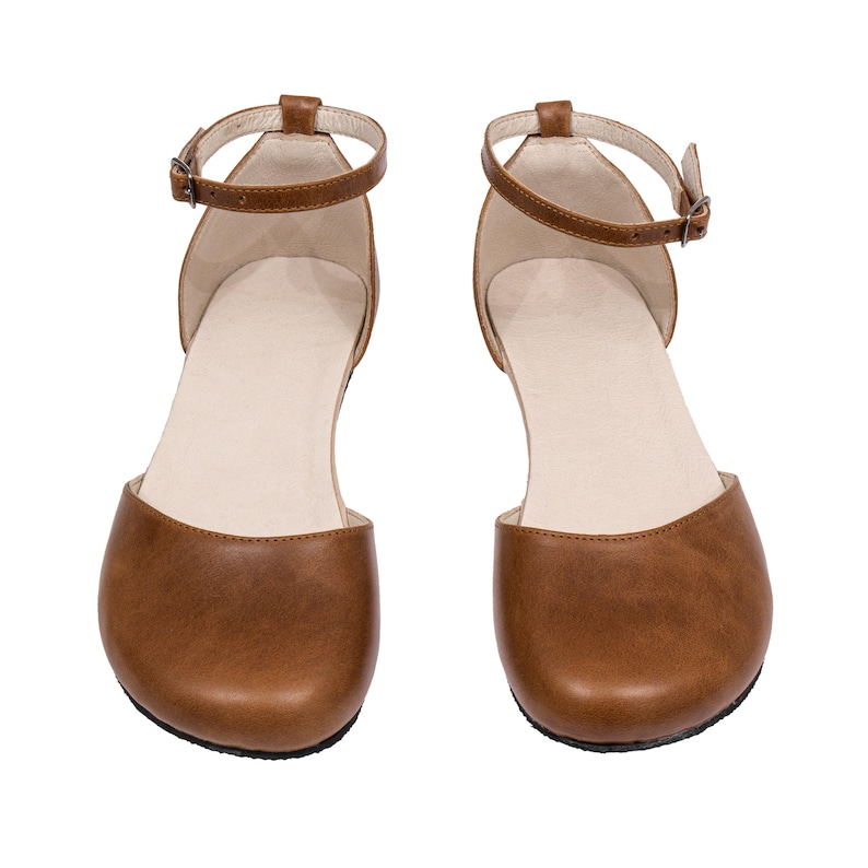 Puede incluir: Un par de sandalias de cuero marr&oacute;n con correas en el tobillo y peque&ntilde;as hebillas. Las sandalias tienen un dise&ntilde;o de punta redondeada y una plantilla de color neutro. Los zapatos se muestran sobre un fondo blanco.