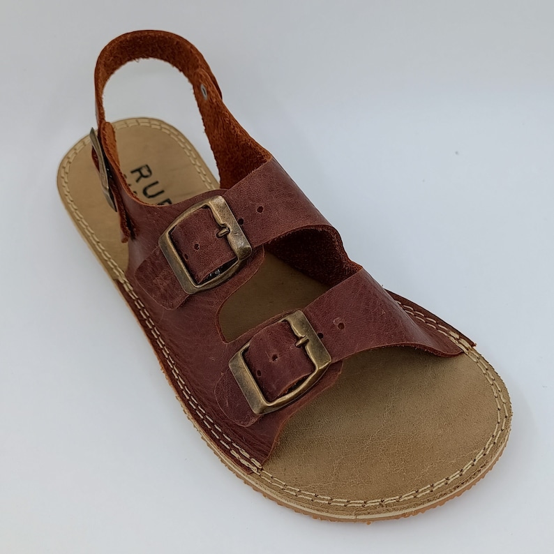 May include: Brown leather sandals with two adjustable buckles and a back strap. The sandals have a light brown sole with a stitched edge.