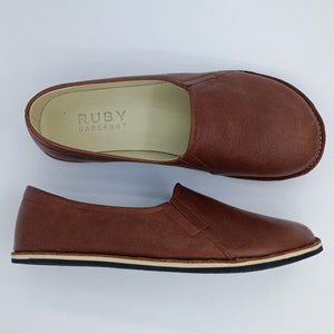 Op de afbeelding: Een paar bruine leren instapschoenen met een witte rubberen zool. De schoenen hebben de tekst "RUBY BAREFOOT" op de binnenzool.