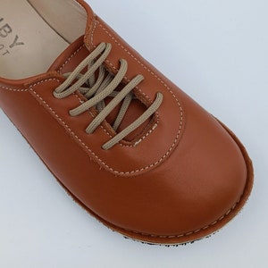 May include: Close-up of a brown leather lace-up shoe with beige laces. The shoe has a stitched seam around the toe and a textured sole.