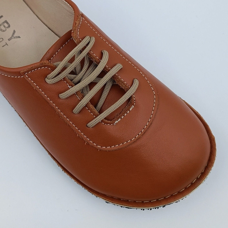 May include: Close-up of a brown leather lace-up shoe with beige laces. The shoe has a stitched seam around the toe and a textured sole.