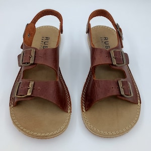 May include: A pair of brown leather sandals with two adjustable buckles. The sandals have a tan leather sole and are labeled "RUBBAREFO".
