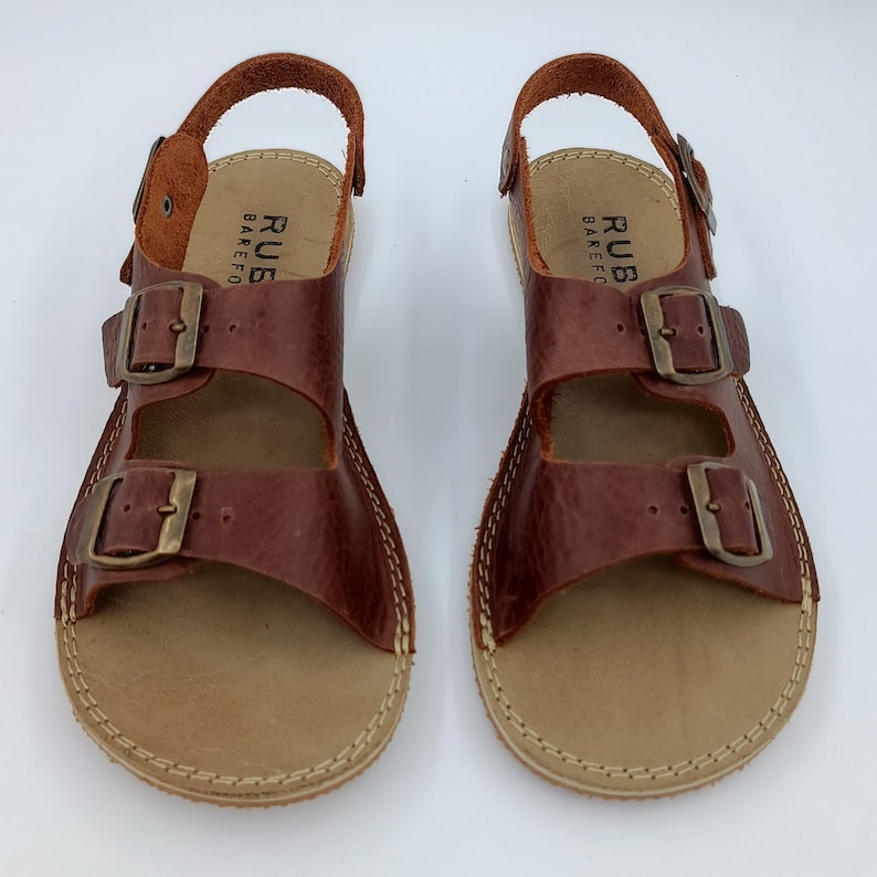 May include: A pair of brown leather sandals with two adjustable buckles. The sandals have a tan leather sole and are labeled "RUBBAREFO".