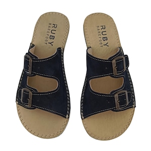 May include: A pair of navy blue sandals with two adjustable straps and metal buckles. The sandals have a beige footbed and the text "RUBY BAREFOOT" is printed on the footbed.