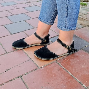 May include: Black suede sandals with ankle straps and a buckle closure. The sandals have a flat sole and are worn on a person's feet.